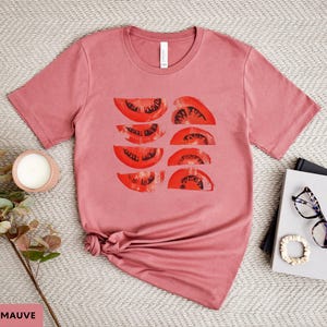 A beautiful, rustic print of eight red ripe tomato slices. Design is multi-color on a Heather Mauve Bella+Canvas 3001 t-shirt. Garment is displayed flat with two books, stylish glasses, candle and decorative eucalyptus branch.