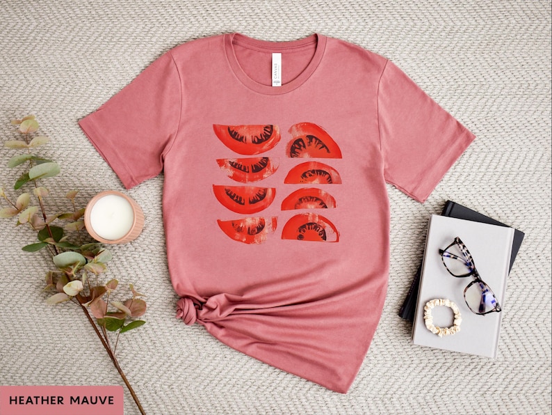 A beautiful, rustic print of eight red ripe tomato slices. Design is multi-color on a Heather Mauve Bella+Canvas 3001 t-shirt. Garment is displayed flat with two books, stylish glasses, candle and decorative eucalyptus branch.