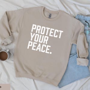 A bold typographic design reading PROTECT YOUR PEACE. Design is white, printed on a Gildan 18000 sweatshirt in Sand colorway. Garment is arranged flat with light blue jeans, glasses and coffee mug.