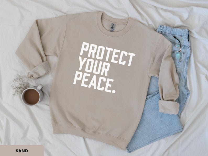 A bold typographic design reading PROTECT YOUR PEACE. Design is white, printed on a Gildan 18000 sweatshirt in Sand colorway. Garment is arranged flat with light blue jeans, glasses and coffee mug.