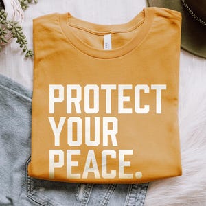 A bold typographic design reading PROTECT YOUR PEACE. Design is white on a Gold Bella+Canvas 3001 t-shirt. Garment is folded with jeans, fedora and succulent plant on a white background.