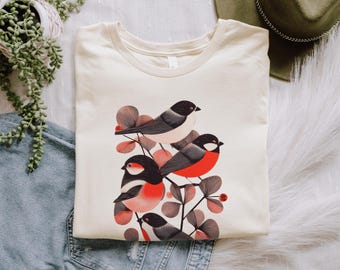 May include: Cream-colored t-shirt featuring a graphic of stylized birds with black heads, gray wings, and red chests. The birds are perched on branches with red and pink leaves. The shirt is folded and displayed on a white surface.