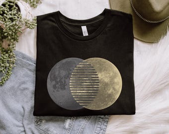 May include: Black t-shirt featuring a graphic design of three overlapping celestial bodies: a gray moon, a gold striped oval, and a yellow moon. The shirt is folded and placed on a white surface with a plant and a hat.