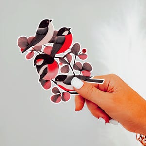 A beautiful and minimal illustration of four finches perched in a tree. Design is black, red and cream, printed on a kiss-cut vinyl sticker with a white border. Sticker is being held by a single hand on a neutral, light grey background.