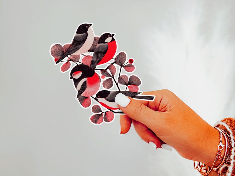 A beautiful and minimal illustration of four finches perched in a tree. Design is black, red and cream, printed on a kiss-cut vinyl sticker with a white border. Sticker is being held by a single hand on a neutral, light grey background.