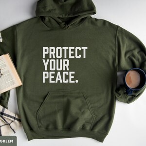 A bold typographic design reading PROTECT YOUR PEACE. Design is white, printed on a Gildan 18500 hoodie in Military Green colorway. Garment is arranged flat with a scarf, book, glasses and coffee mug.
