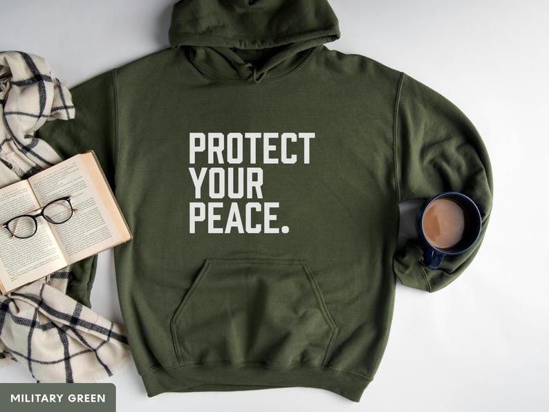 A bold typographic design reading PROTECT YOUR PEACE. Design is white, printed on a Gildan 18500 hoodie in Military Green colorway. Garment is arranged flat with a scarf, book, glasses and coffee mug.