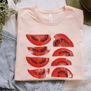 A beautiful, rustic print of eight red ripe tomato slices. Design is multi-color on a Heather Peach Bella+Canvas 3001 t-shirt. Garment is folded with jeans, fedora and succulent plant on a white background.