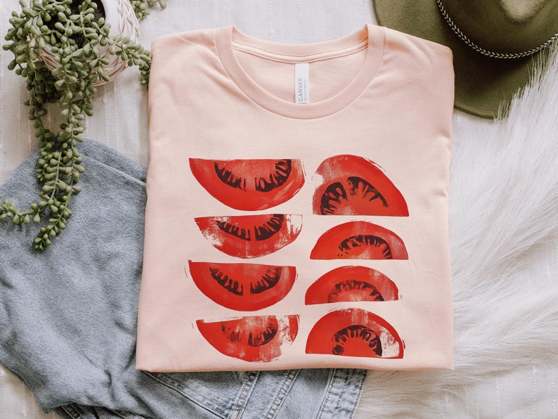 A beautiful, rustic print of eight red ripe tomato slices. Design is multi-color on a Heather Peach Bella+Canvas 3001 t-shirt. Garment is folded with jeans, fedora and succulent plant on a white background.