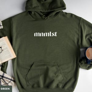 A minimalist typographic design that reads "mnmlst". Design is printed on a Gildan 18500 sweatshirt in Military Green colorway. Garment is arranged flat with light blue jeans, glasses and coffee mug.