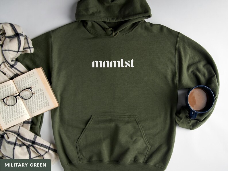 A minimalist typographic design that reads "mnmlst". Design is printed on a Gildan 18500 sweatshirt in Military Green colorway. Garment is arranged flat with light blue jeans, glasses and coffee mug.