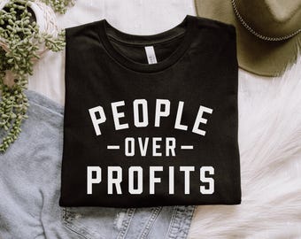 May include: Black t-shirt with the white text "PEOPLE OVER PROFITS". The shirt is folded and placed on a white fluffy surface, with a pair of blue jeans and a plant in the background.
