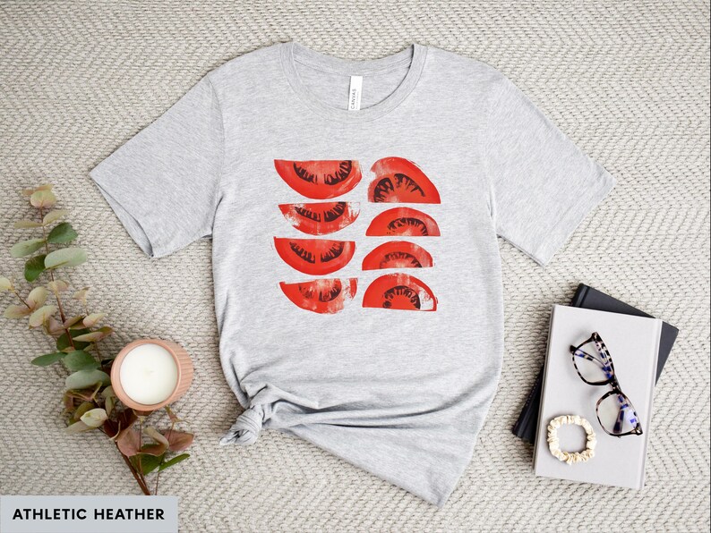 A beautiful, rustic print of eight red ripe tomato slices. Design is multi-color on a Athletic Heather Grey Bella+Canvas 3001 t-shirt. Garment is displayed flat with two books, stylish glasses, candle and decorative eucalyptus branch.