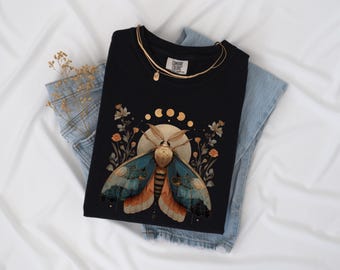 May include: A black t-shirt featuring a graphic print of a large moth with blue and orange wings, positioned in front of a full moon. Above the moth are golden moon phases and floral elements. The shirt is layered over light blue denim jeans.