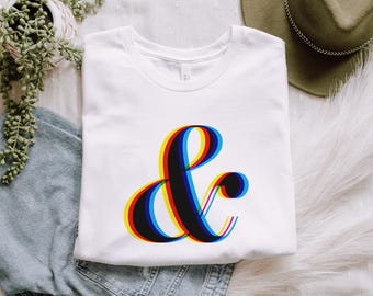 May include: White t-shirt featuring a large, stylized ampersand (&) graphic. The ampersand has a 3D effect with red, yellow, and blue outlines. The shirt is folded and placed on a white surface, with a pair of jeans and a decorative plant visible.