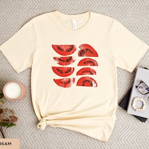 A beautiful, rustic print of eight red ripe tomato slices. Design is multi-color on a Soft Cream Bella+Canvas 3001 t-shirt. Garment is displayed flat with two books, stylish glasses, candle and decorative eucalyptus branch.