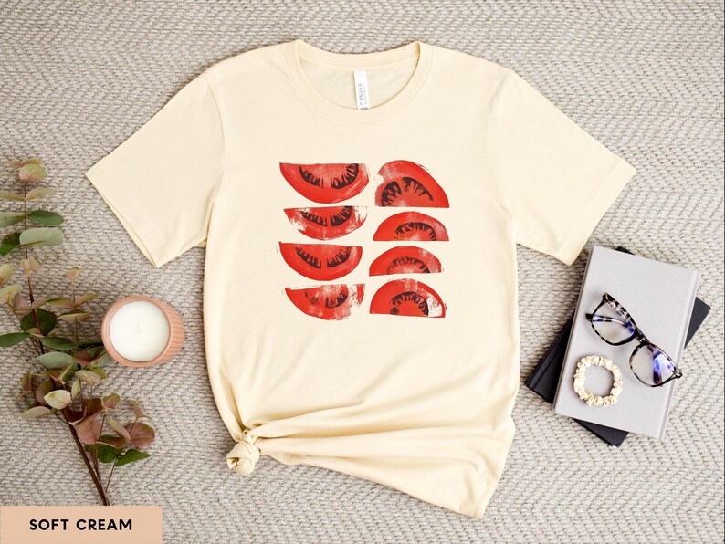 A beautiful, rustic print of eight red ripe tomato slices. Design is multi-color on a Soft Cream Bella+Canvas 3001 t-shirt. Garment is displayed flat with two books, stylish glasses, candle and decorative eucalyptus branch.