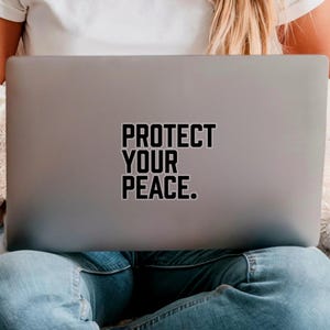 A bold typographic design reading PROTECT YOUR PEACE. Design is printed on a kiss-cut vinyl sticker with a white border. Sticker is shown on the back of an open silver laptop as a young lady is working.