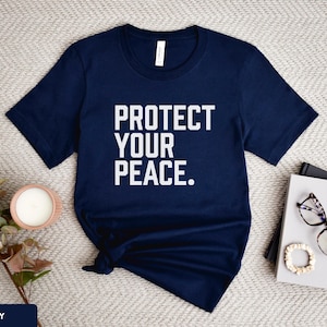 A bold typographic design reading PROTECT YOUR PEACE. Design is white on a navy Bella+Canvas 3001 t-shirt. Garment is displayed flat with jeans, two books, candle and decorative eucalyptus branch.