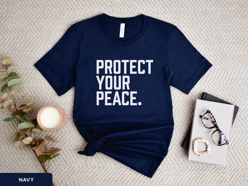 A bold typographic design reading PROTECT YOUR PEACE. Design is white on a navy Bella+Canvas 3001 t-shirt. Garment is displayed flat with jeans, two books, candle and decorative eucalyptus branch.
