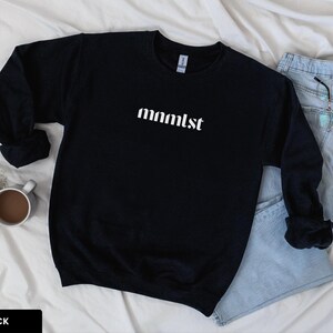 A minimalist typographic design that reads "mnmlst". Design is printed on a Gildan 18000 sweatshirt in Black colorway. Garment is arranged flat with light blue jeans, glasses and coffee mug.