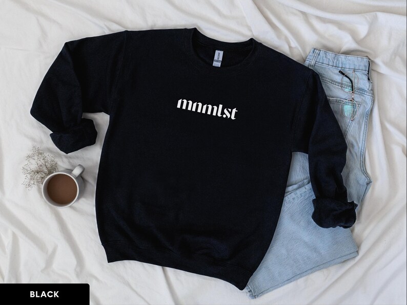 A minimalist typographic design that reads "mnmlst". Design is printed on a Gildan 18000 sweatshirt in Black colorway. Garment is arranged flat with light blue jeans, glasses and coffee mug.