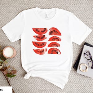 A beautiful, rustic print of eight red ripe tomato slices. Design is multi-color on a White Bella+Canvas 3001 t-shirt. Garment is displayed flat with two books, stylish glasses, candle and decorative eucalyptus branch.