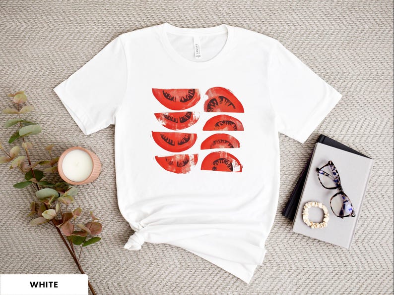 A beautiful, rustic print of eight red ripe tomato slices. Design is multi-color on a White Bella+Canvas 3001 t-shirt. Garment is displayed flat with two books, stylish glasses, candle and decorative eucalyptus branch.