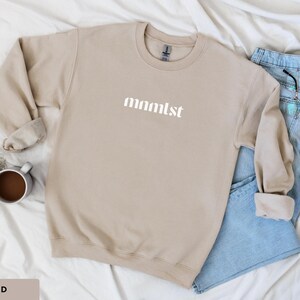 A minimalist typographic design that reads "mnmlst". Design is printed on a Gildan 18000 sweatshirt in Sand colorway. Garment is arranged flat with light blue jeans, glasses and coffee mug.