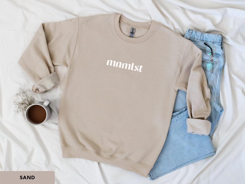 A minimalist typographic design that reads "mnmlst". Design is printed on a Gildan 18000 sweatshirt in Sand colorway. Garment is arranged flat with light blue jeans, glasses and coffee mug.