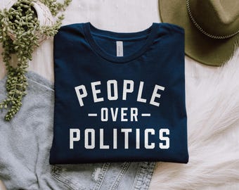 May include: Navy blue crew neck t-shirt with the white text "PEOPLE OVER POLITICS". The shirt is folded and placed on a white fur-like surface, with a green hat and a plant in the background.