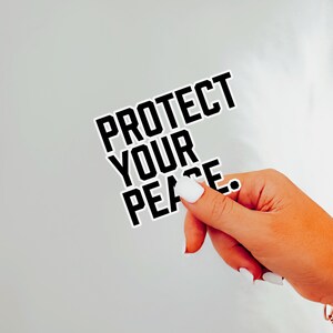 A bold typographic design reading PROTECT YOUR PEACE. Design is printed on a kiss-cut vinyl sticker with a white border. Sticker is being held by a single hand on a neutral, light grey background.
