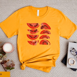 A beautiful, rustic print of eight red ripe tomato slices. Design is multi-color on a Gold Bella+Canvas 3001 t-shirt. Garment is displayed flat with two books, stylish glasses, candle and decorative eucalyptus branch.
