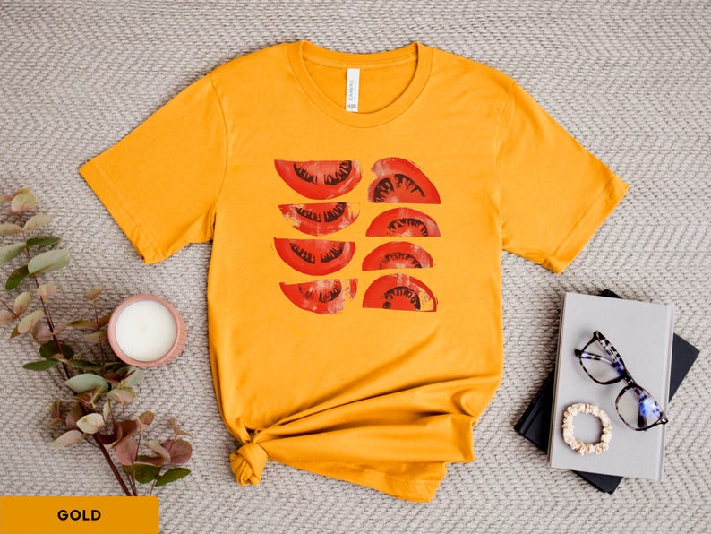 A beautiful, rustic print of eight red ripe tomato slices. Design is multi-color on a Gold Bella+Canvas 3001 t-shirt. Garment is displayed flat with two books, stylish glasses, candle and decorative eucalyptus branch.