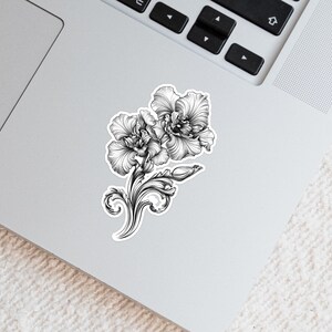 An ornate engraved style rendering of two orchids with elegant flourishes in a balanced, elegant arrangement. Design is printed on a kiss-cut vinyl sticker with a white border. Sticker is shown on an open silver laptop, next to the track pad.