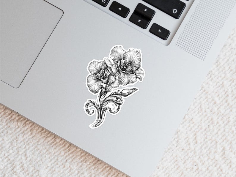 An ornate engraved style rendering of two orchids with elegant flourishes in a balanced, elegant arrangement. Design is printed on a kiss-cut vinyl sticker with a white border. Sticker is shown on an open silver laptop, next to the track pad.