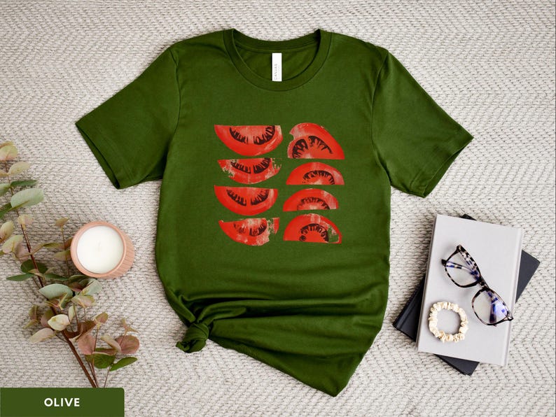 A beautiful, rustic print of eight red ripe tomato slices. Design is multi-color on a Olive Bella+Canvas 3001 t-shirt. Garment is displayed flat with two books, stylish glasses, candle and decorative eucalyptus branch.