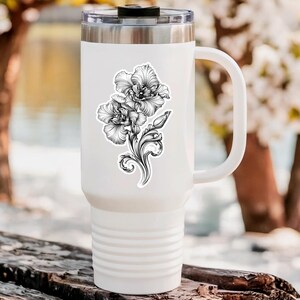 An ornate engraved style rendering of two orchids with elegant flourishes in a balanced, elegant arrangement. Design is printed on a kiss-cut vinyl sticker with a white border. Sticker is shown on a tall white tumbler in a luxurious outdoor setting.