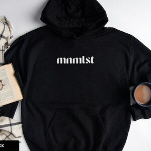 A minimalist typographic design that reads "mnmlst". Design is printed on a Gildan 18500 sweatshirt in Black colorway. Garment is arranged flat with light blue jeans, glasses and coffee mug.