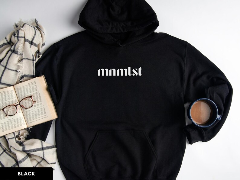 A minimalist typographic design that reads "mnmlst". Design is printed on a Gildan 18500 sweatshirt in Black colorway. Garment is arranged flat with light blue jeans, glasses and coffee mug.
