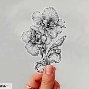 An ornate engraved style rendering of two orchids with elegant flourishes in a balanced, elegant arrangement. Design is printed on a kiss-cut vinyl sticker with a transparent border. Sticker is being held by a single hand on a neutral, light grey background.
