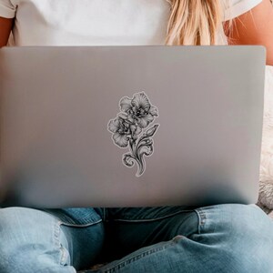 An ornate engraved style rendering of two orchids with elegant flourishes in a balanced, elegant arrangement. Design is printed on a kiss-cut vinyl sticker with a white border. Sticker is shown on the back of an open silver laptop as a young lady is working.