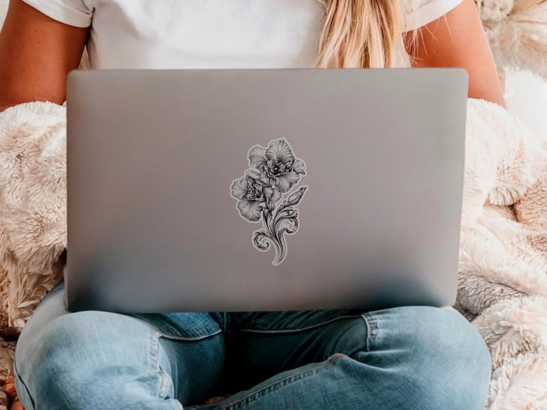 An ornate engraved style rendering of two orchids with elegant flourishes in a balanced, elegant arrangement. Design is printed on a kiss-cut vinyl sticker with a white border. Sticker is shown on the back of an open silver laptop as a young lady is working.