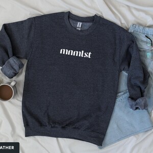 A minimalist typographic design that reads "mnmlst". Design is printed on a Gildan 18000 sweatshirt in Dark Heather colorway. Garment is arranged flat with light blue jeans, glasses and coffee mug.