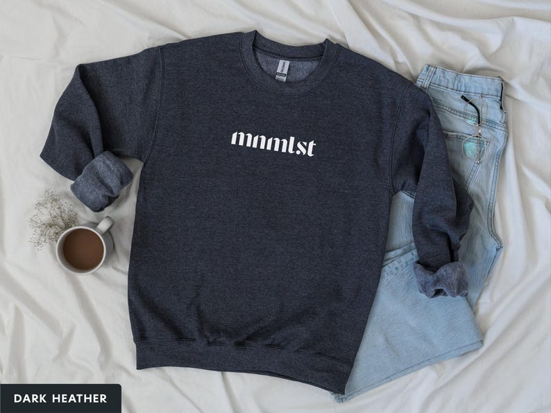 A minimalist typographic design that reads "mnmlst". Design is printed on a Gildan 18000 sweatshirt in Dark Heather colorway. Garment is arranged flat with light blue jeans, glasses and coffee mug.