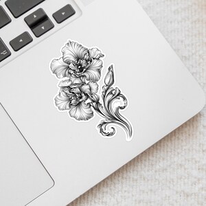 An ornate engraved style rendering of two orchids with elegant flourishes in a balanced, elegant arrangement. Design is printed on a kiss-cut vinyl sticker with a white border. Sticker is shown on an open silver laptop, next to the track pad.