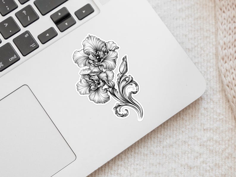 An ornate engraved style rendering of two orchids with elegant flourishes in a balanced, elegant arrangement. Design is printed on a kiss-cut vinyl sticker with a white border. Sticker is shown on an open silver laptop, next to the track pad.