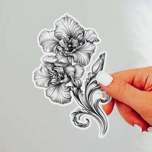 An ornate engraved style rendering of two orchids with elegant flourishes in a balanced, elegant arrangement. Design is printed on a kiss-cut vinyl sticker with a white border. Sticker is being held by a single hand on a neutral, light grey background.