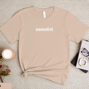A minimalist typographic design that reads "mnmlst". Design is white on an Sand Bella+Canvas 3001 t-shirt. Garment is displayed flat with two books, stylish glasses, candle and decorative eucalyptus branch.