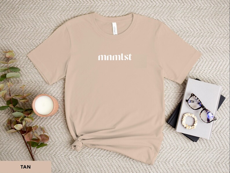 A minimalist typographic design that reads "mnmlst". Design is white on an Sand Bella+Canvas 3001 t-shirt. Garment is displayed flat with two books, stylish glasses, candle and decorative eucalyptus branch.